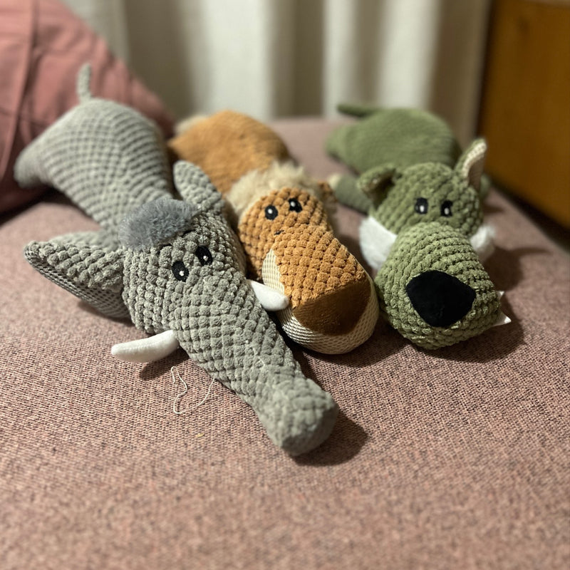 Trois peluches d'animaux en tissu texturé sur un canapé rose : un éléphant gris, un renard orange et un chien vert. Jouets doux pour enfants.