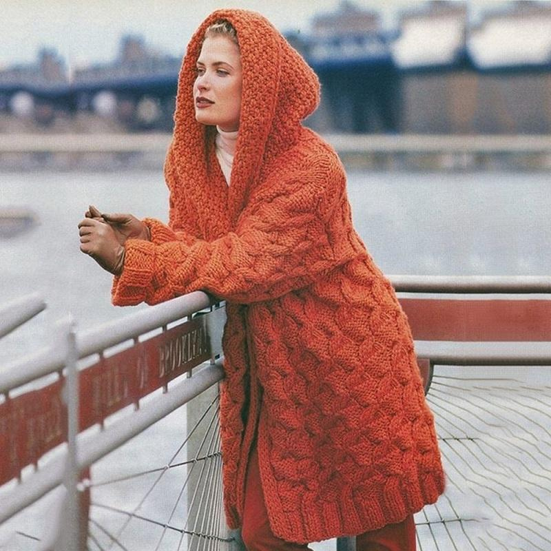 Femme portant un manteau en laine orange avec capuche, debout près d'une balustrade, fond urbain flou. Mode hiver, tricot tendance.