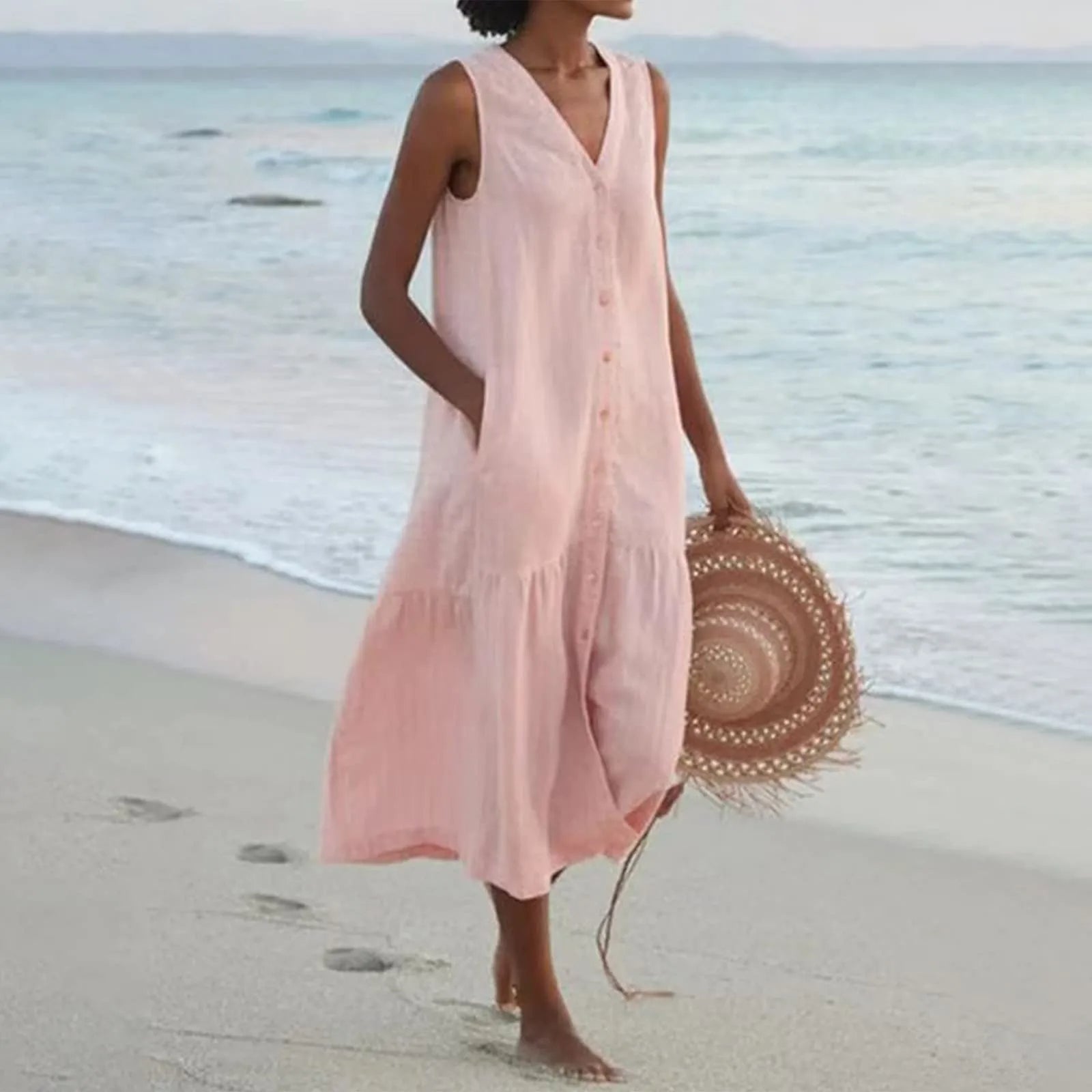 Robe d'été rose sans manches sur plage, femme tenant un chapeau rond en paille, mer en arrière-plan, mode estivale élégante et décontractée.