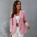Femme souriante portant un blazer rose élégant, top blanc et pantalon blanc, posant contre un mur gris. Mode féminine, tenue chic, style moderne.
