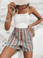 Femme en débardeur blanc et short rayé multicolore, posant contre un mur blanc. Mode estivale, tenue décontractée, accessoires élégants.