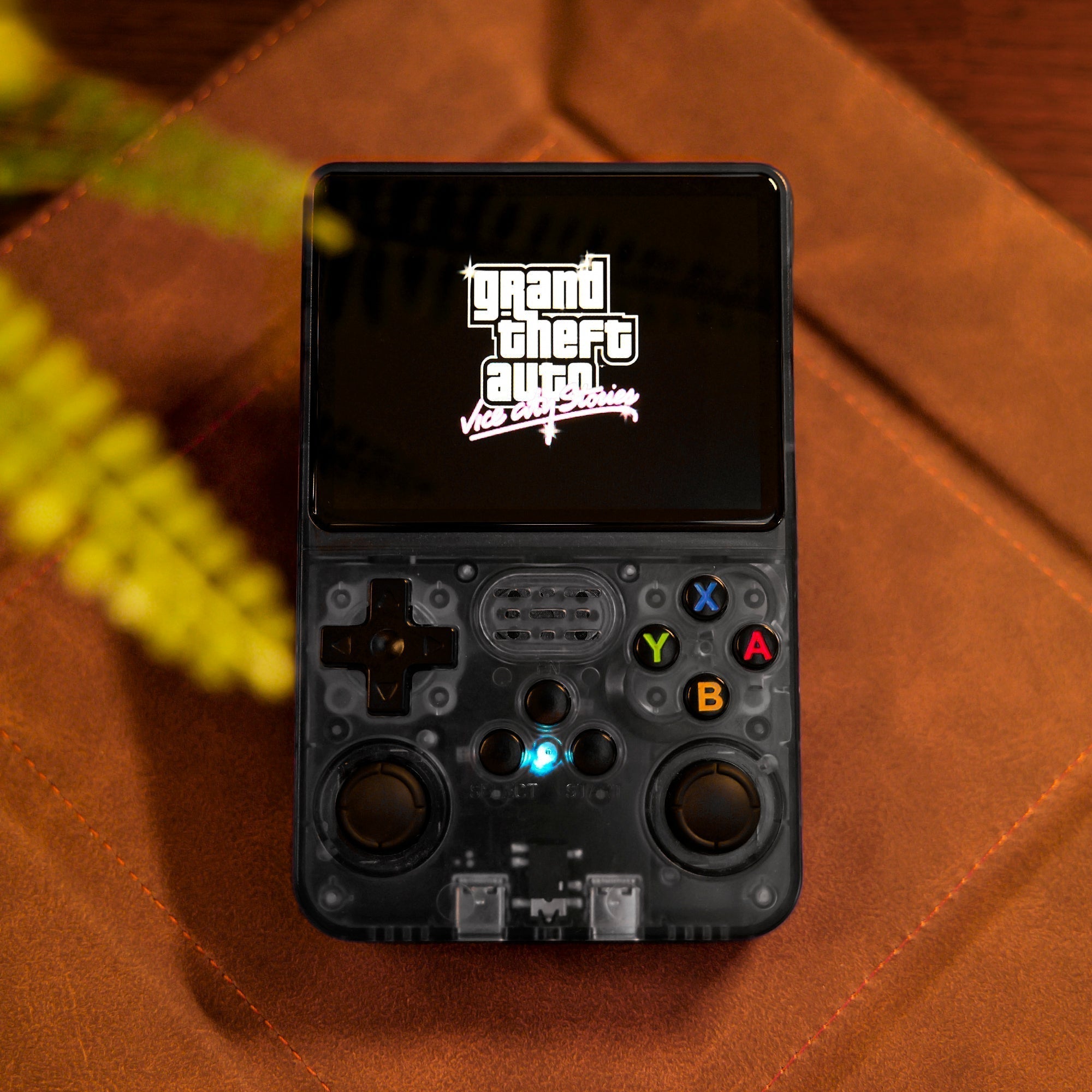 Console de jeu portable rétro avec écran affichant "Grand Theft Auto". Design compact, boutons colorés, sur fond de cuir marron. Accessoire gaming tendance.