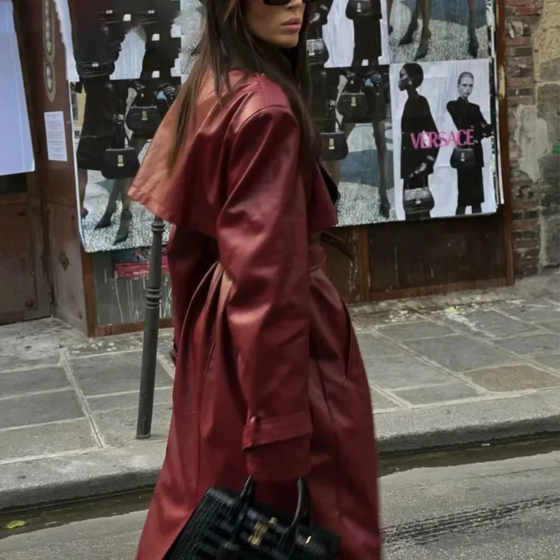 Femme portant un long manteau en cuir rouge, lunettes de soleil noires, sac à main noir, marchant dans une rue urbaine avec affiches en arrière-plan.