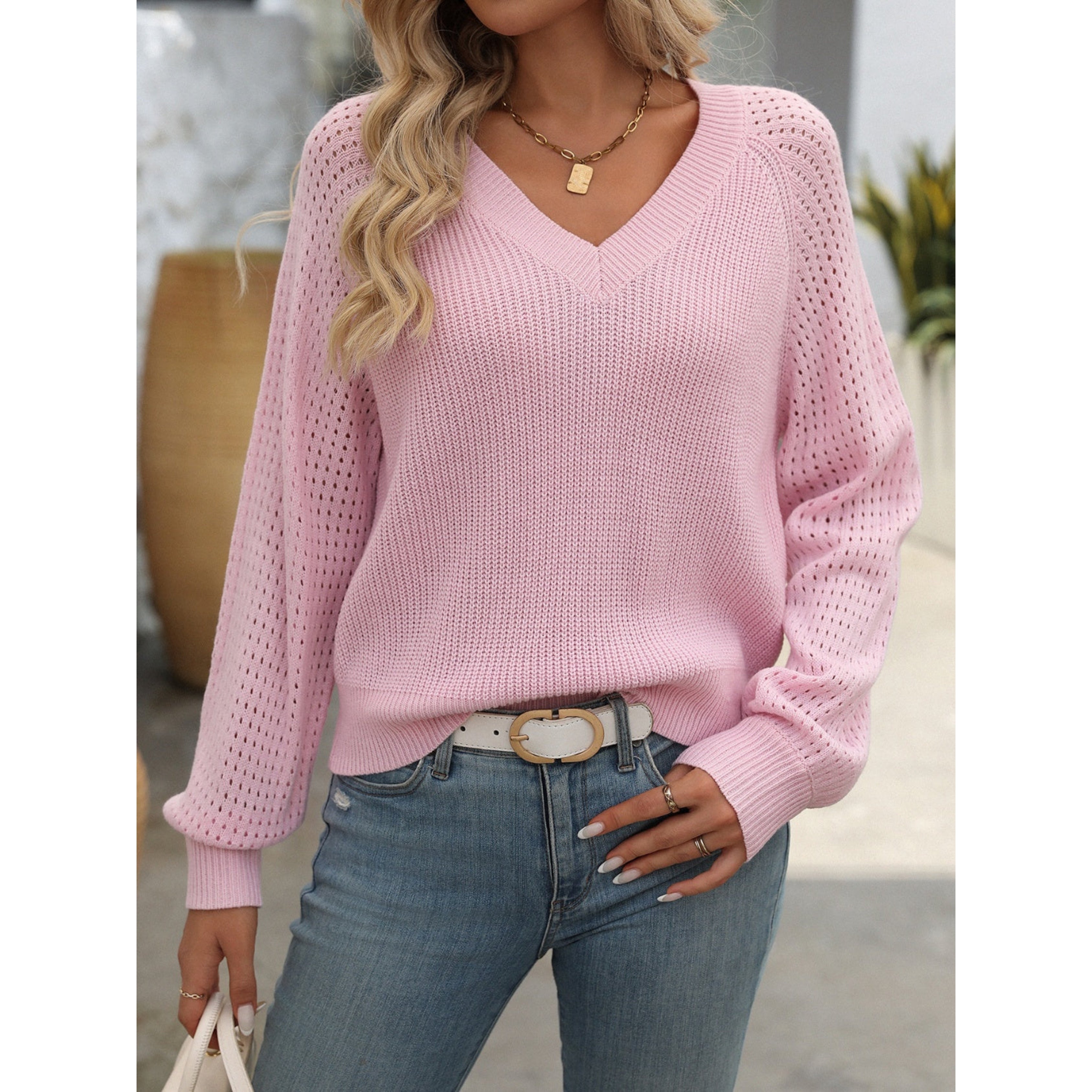 Femme portant un pull rose en maille ajourée avec un col en V, associé à un jean bleu et une ceinture blanche. Mode féminine décontractée.