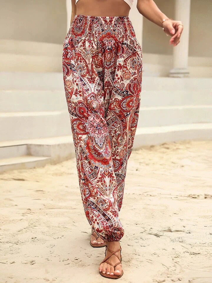 Pantalon ample à motifs cachemire rouge et blanc, porté avec des sandales marron sur fond de plage. Mode estivale, style bohème chic.