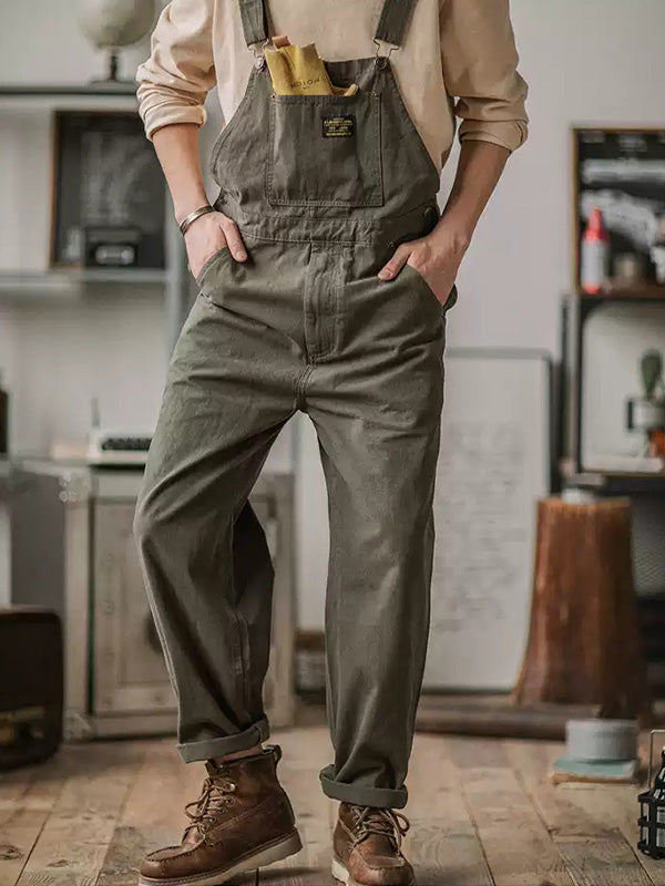 Homme portant une salopette verte avec un t-shirt beige, mains dans les poches, debout dans un intérieur rustique. Mode décontractée, style vintage.