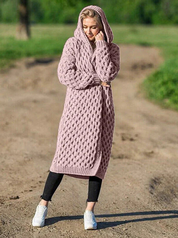 Femme portant un long cardigan en tricot rose avec capuche, debout sur un chemin de terre, style décontracté, mode automne-hiver, confort et élégance.