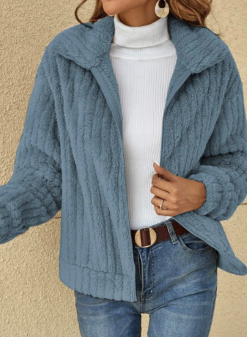 Femme portant une veste en fausse fourrure bleue, col montant, sur un pull blanc à col roulé, avec un jean bleu. Mode automne-hiver élégante.