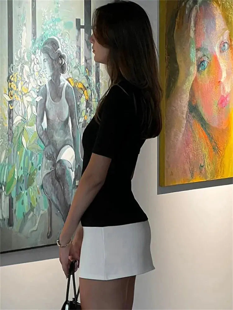 Femme observant des tableaux dans une galerie d'art moderne, portant une jupe blanche et un haut noir, éclairage doux, ambiance contemplative.