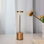 Lampe de table LED dorée moderne sur un bureau blanc, éclairant une pièce minimaliste avec rideaux blancs et vase de fleurs, design élégant et contemporain.