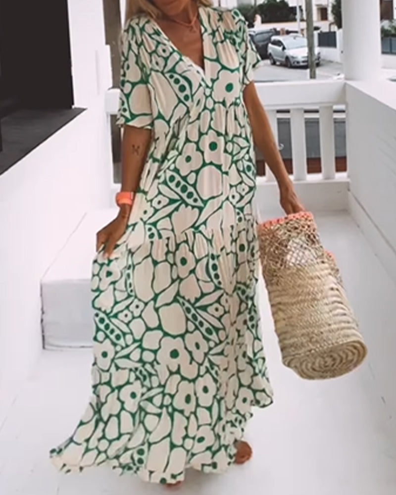 Robe longue à motifs floraux verts et blancs, portée par une femme sur une terrasse. Elle tient un sac en osier. Mode estivale élégante et décontractée.