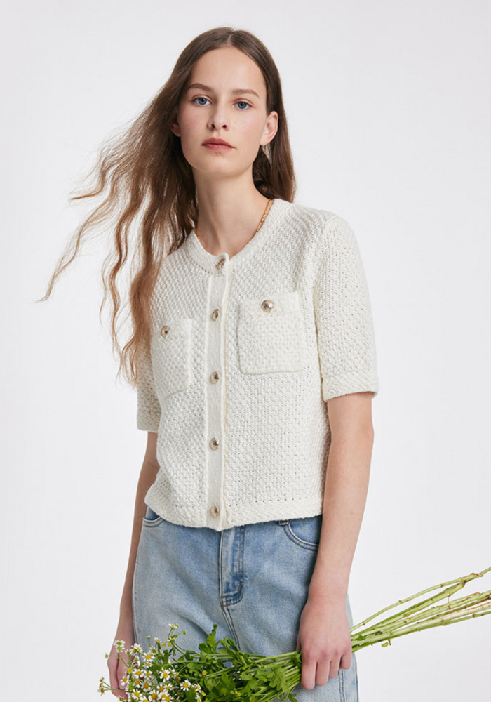 Femme portant un cardigan blanc en tricot avec boutons dorés, poches avant, manches courtes, tenant un bouquet de fleurs, mode décontractée élégante.