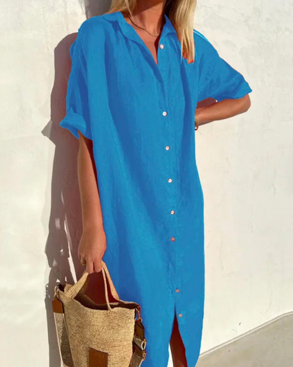 Robe chemise bleue en lin portée par une femme, manches courtes, avec sac en paille, contre un mur blanc. Mode estivale, tenue décontractée.