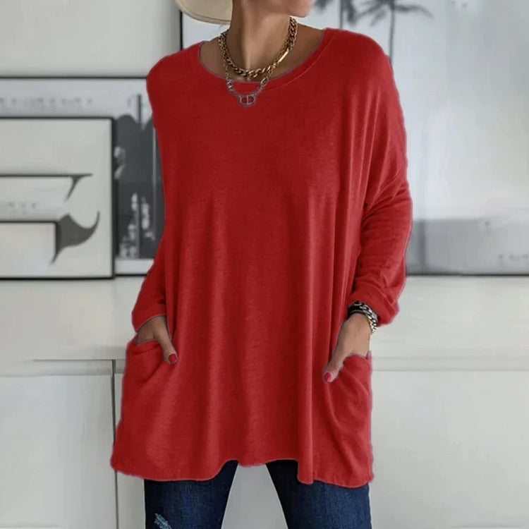 Femme portant un pull rouge ample avec poches, collier doré, chapeau beige, posant devant un mur blanc. Mode décontractée, tendance automne.