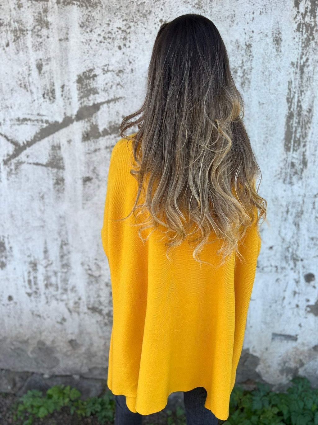 Femme de dos avec cheveux longs ondulés, portant un poncho jaune vif, devant un mur texturé. Mode automne, style décontracté, tendance cheveux.