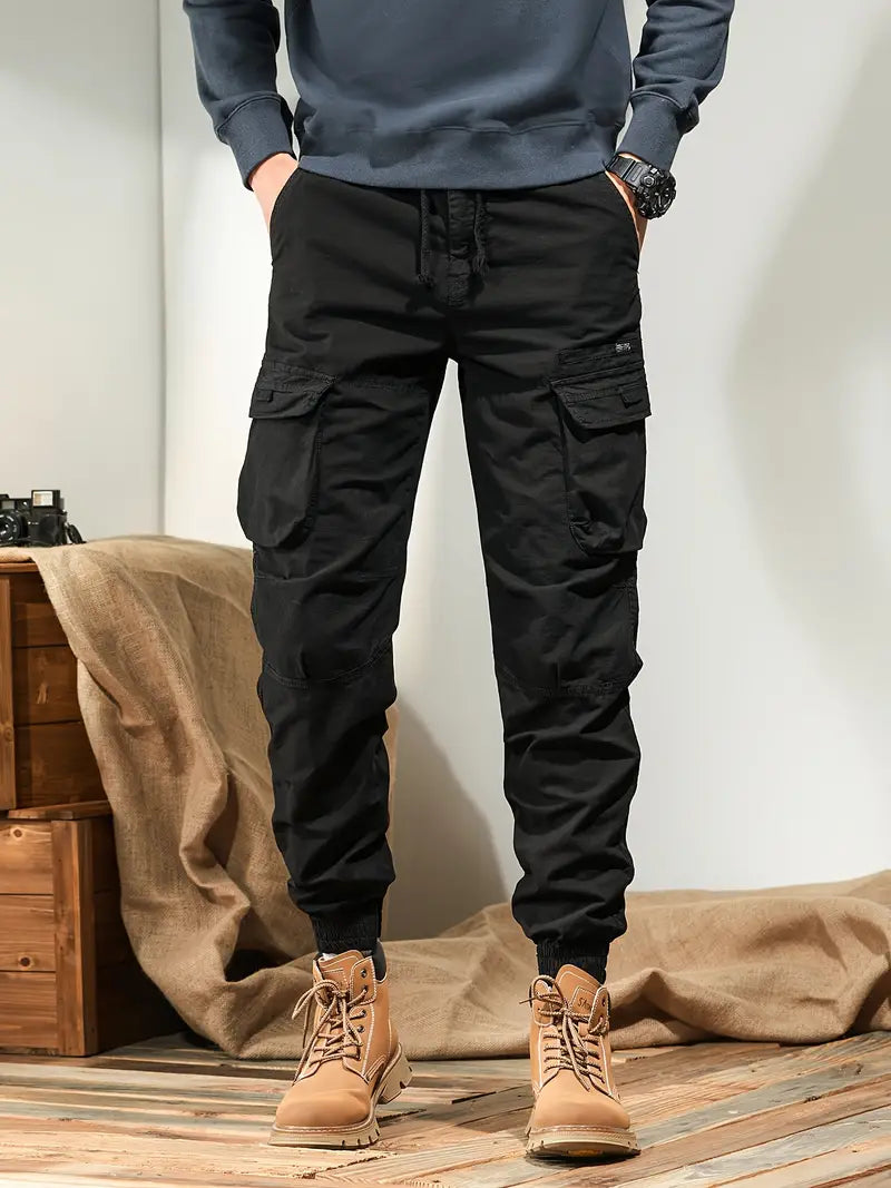 Pantalon cargo noir pour homme avec poches multiples, style décontracté, associé à des bottes marron, sur fond intérieur moderne.