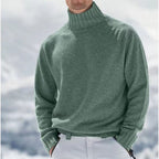 Homme portant un pull col roulé vert en laine, style hiver, devant un paysage enneigé. Mode masculine, vêtements chauds, tendance hiver.