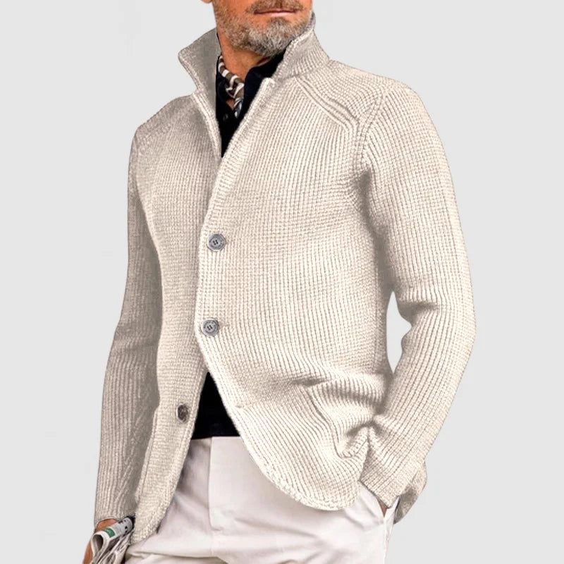 Homme portant un cardigan beige élégant, col relevé, boutons visibles, sur chemise noire et pantalon clair. Mode masculine chic et moderne.
