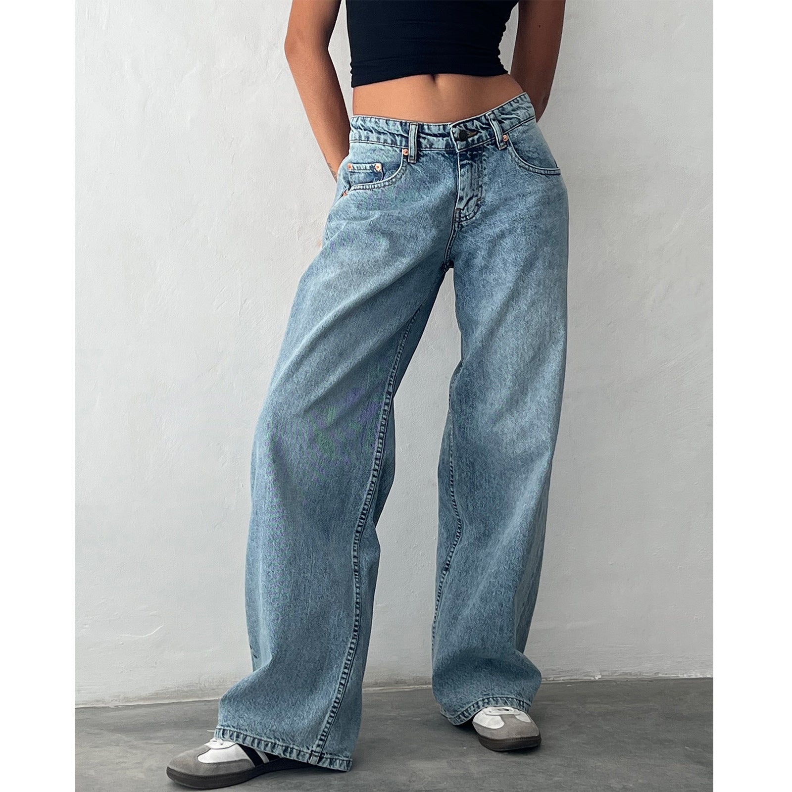 Jeans larges en denim bleu clair portés par une personne debout, associés à un haut noir court et des baskets grises, style décontracté moderne.