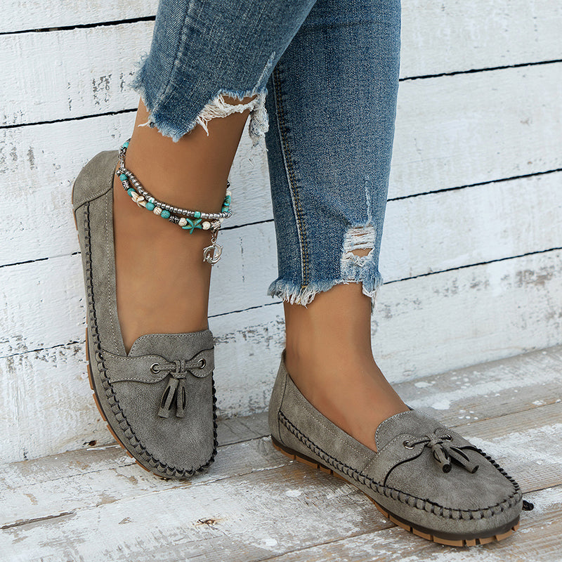 Chaussures mocassins gris en daim avec nœuds, portées avec un jean déchiré et bracelet de cheville turquoise, posées sur fond de bois blanc.