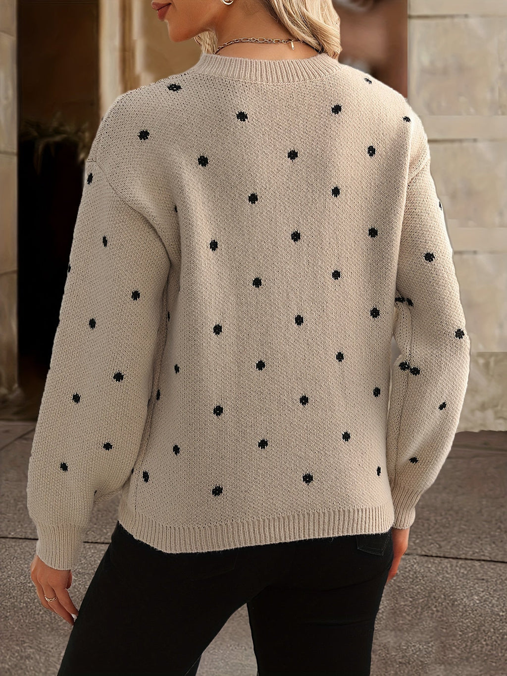 Femme portant un pull beige à pois noirs, manches longues, col rond, style décontracté, mode automne-hiver, arrière-plan urbain flou.
