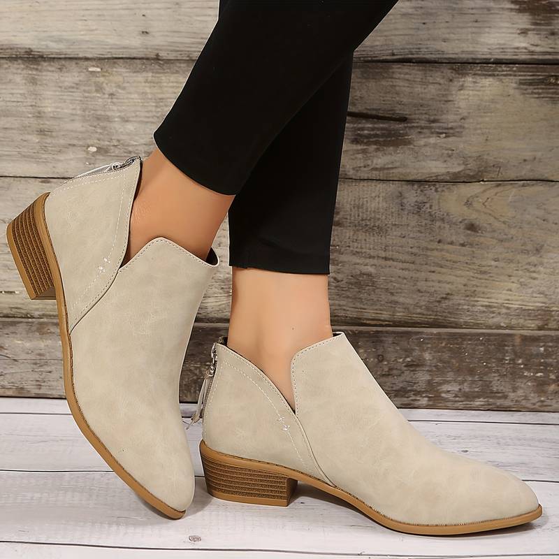 Bottines beiges en cuir synthétique, talon bas, portées avec un pantalon noir, posées sur un sol en bois. Mode féminine, chaussures tendance.