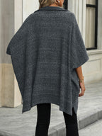 Femme portant un poncho gris en tricot, style décontracté, devant un bâtiment moderne. Mode automne-hiver, tenue élégante et confortable.