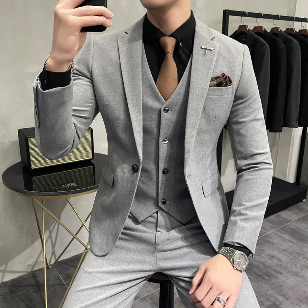 Homme en costume gris élégant, cravate marron, chemise noire, montre argentée, assis, mode masculine, style formel, boutique de vêtements.