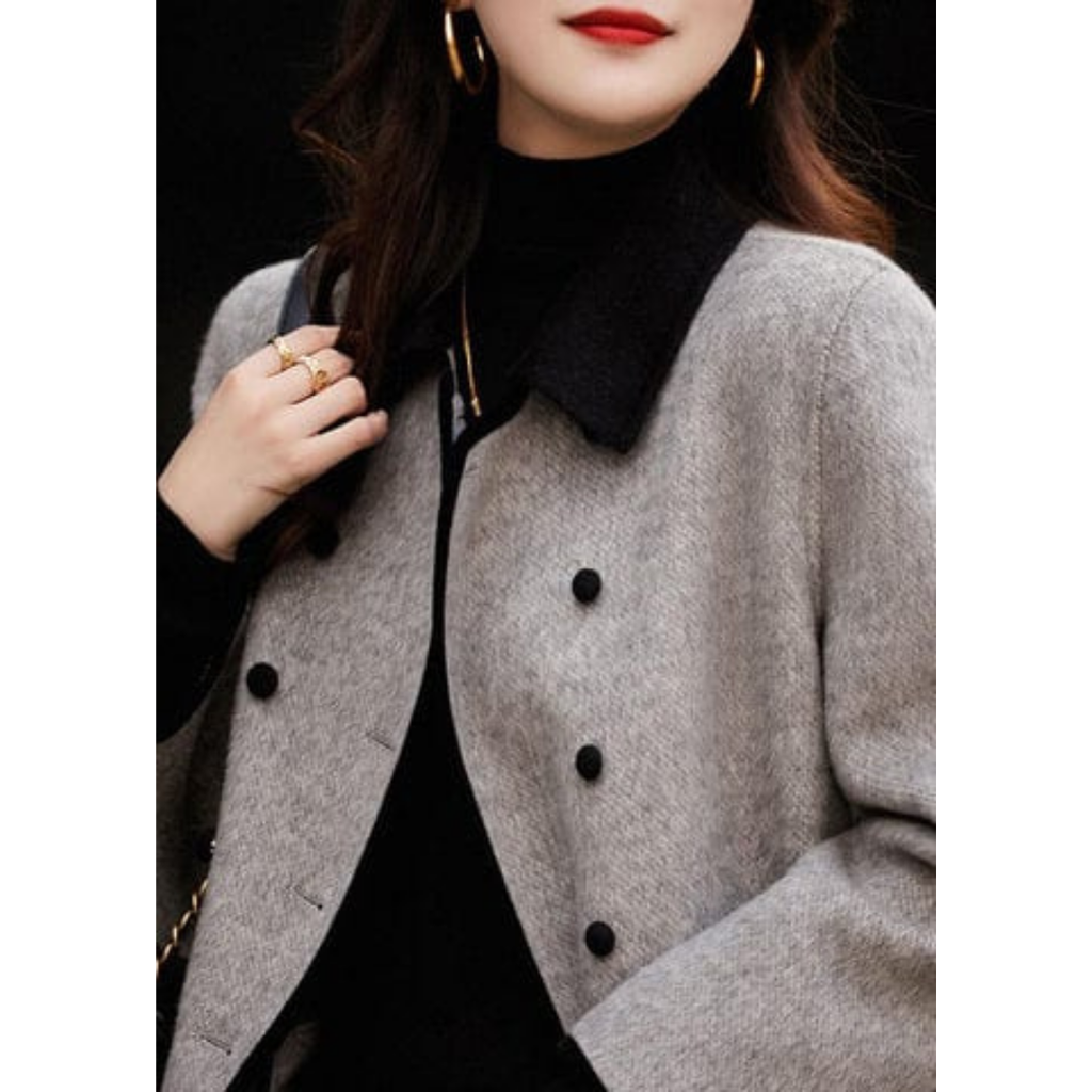 Femme élégante portant un manteau gris à double boutonnage, col noir, avec pull noir et boucles d'oreilles dorées. Mode automne chic.