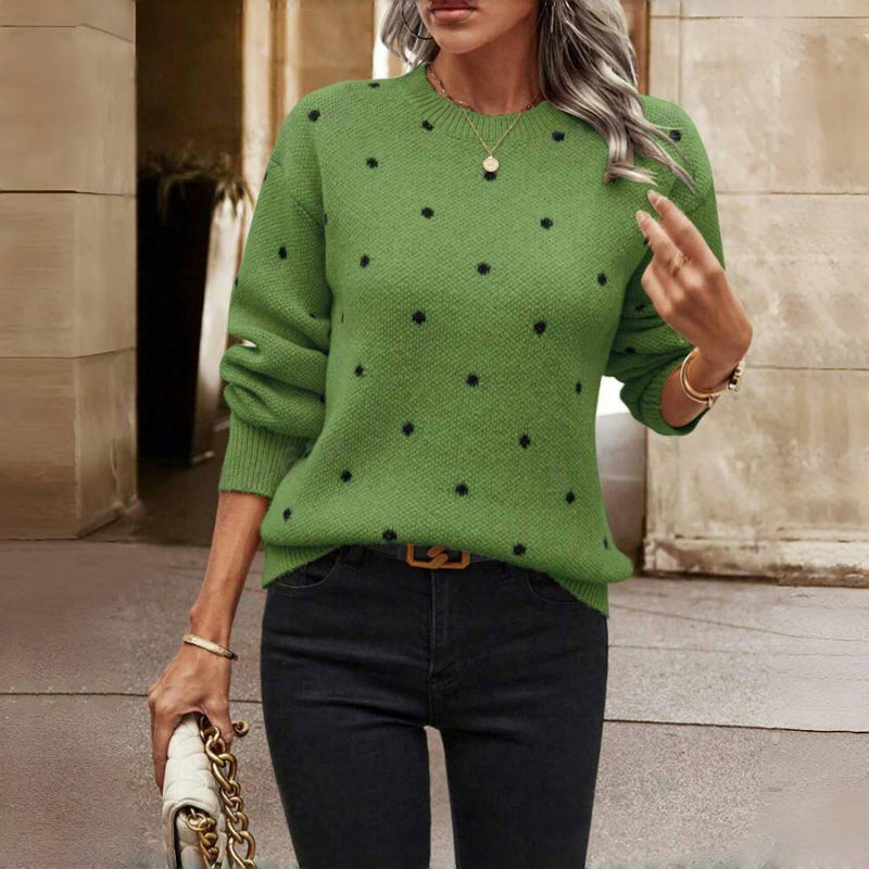 Femme portant un pull vert à pois noirs, tenant un sac à main blanc avec chaîne dorée, debout dans une rue urbaine. Mode automne chic et élégante.