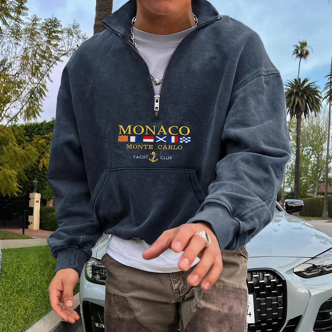 Homme portant un sweat-shirt bleu marine "Monaco Monte Carlo Yacht Club" devant une voiture de luxe, entouré de palmiers et d'arbres.