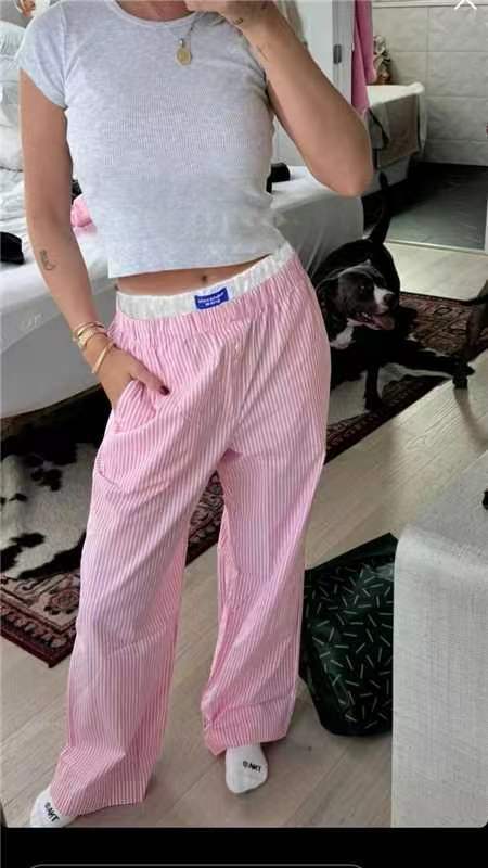 Femme en t-shirt blanc et pantalon rose à rayures, posant dans une chambre moderne avec un chien noir et blanc en arrière-plan.