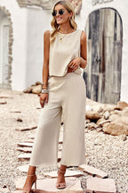 Femme en tenue beige élégante, lunettes de soleil, posant sur un chemin de pierres. Mode estivale chic, combinaison sans manches, sandales à talons.