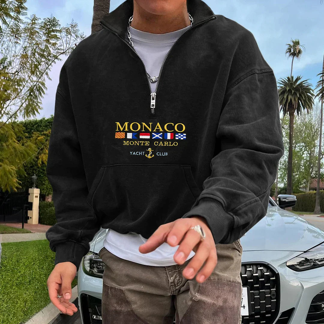 Homme portant un sweat noir "Monaco Yacht Club" devant une voiture de luxe, entouré de palmiers. Mode décontractée, style urbain chic.