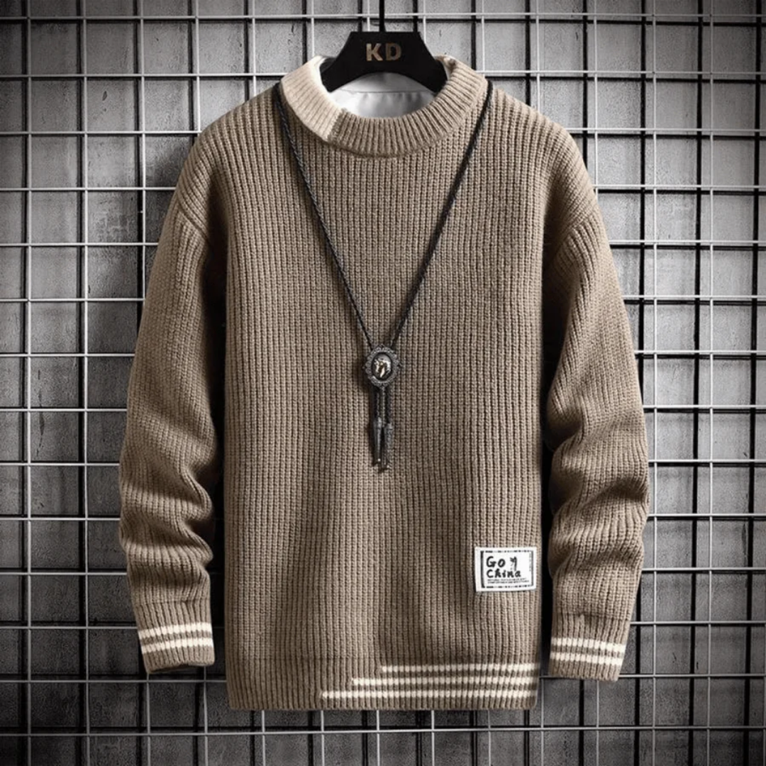 Pull beige en tricot côtelé avec collier pendentif, accroché sur un cintre noir devant un mur à carreaux gris. Mode automne-hiver tendance.