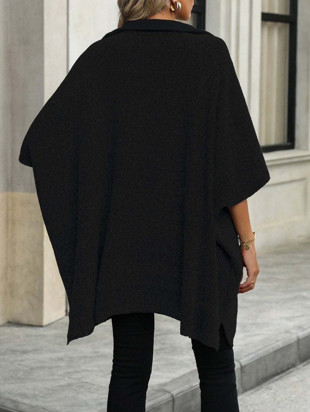 Femme portant un poncho noir élégant, debout devant un bâtiment moderne. Mode automne, tenue chic, style urbain.
