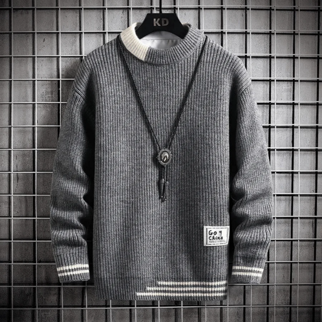 Pull gris en tricot épais avec col rond, détails blancs aux poignets et col, pendentif noir, accroché sur un cintre devant un mur grillagé.