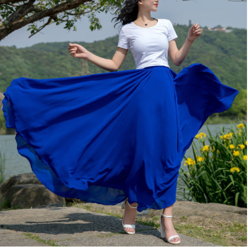 Femme en jupe longue bleue virevoltante, t-shirt blanc, posant près d'un lac et de fleurs jaunes, style estival, mode féminine élégante.