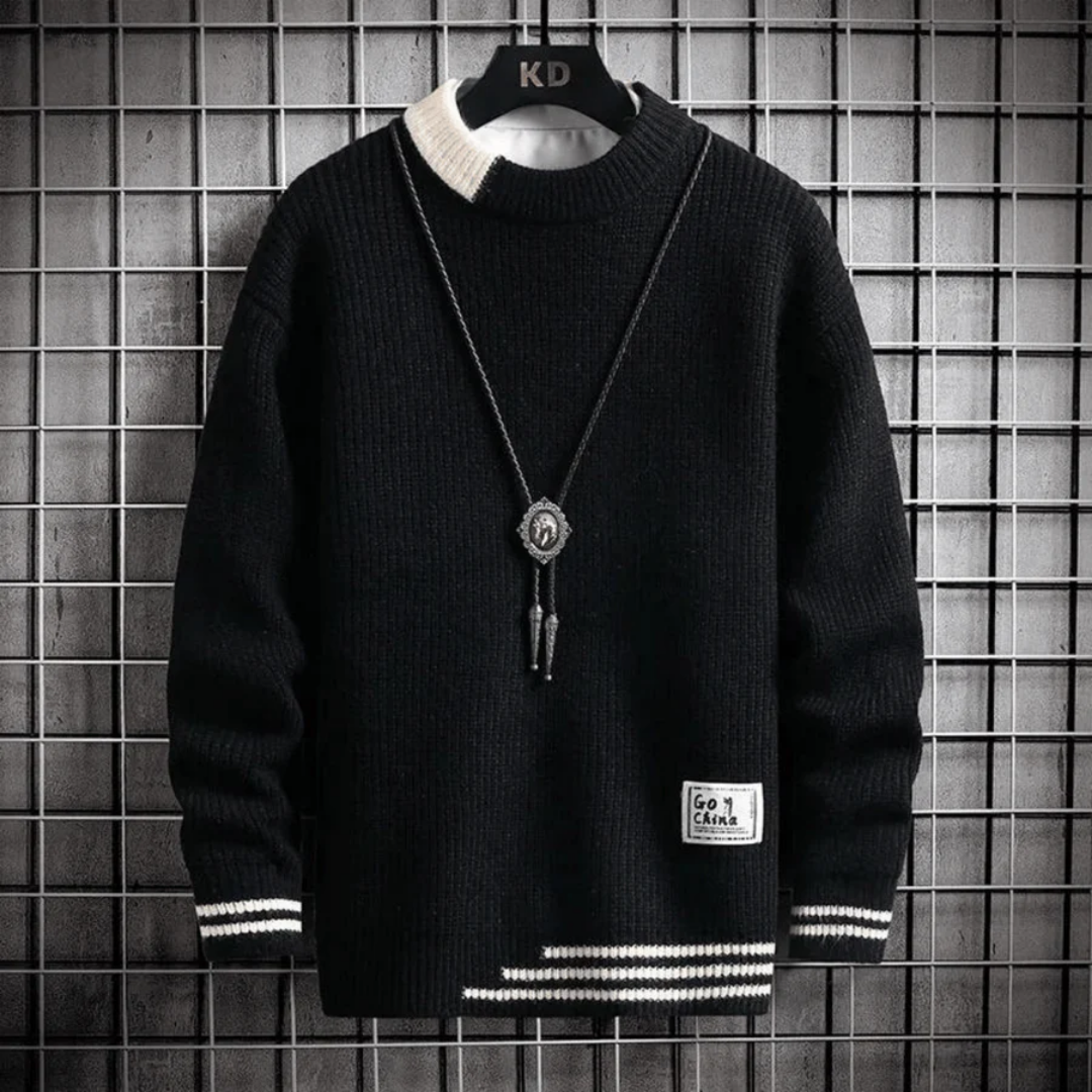 Pull noir en tricot épais avec col rond, détails blancs aux poignets et à l'ourlet, pendentif argenté, accroché sur un fond de grille métallique.