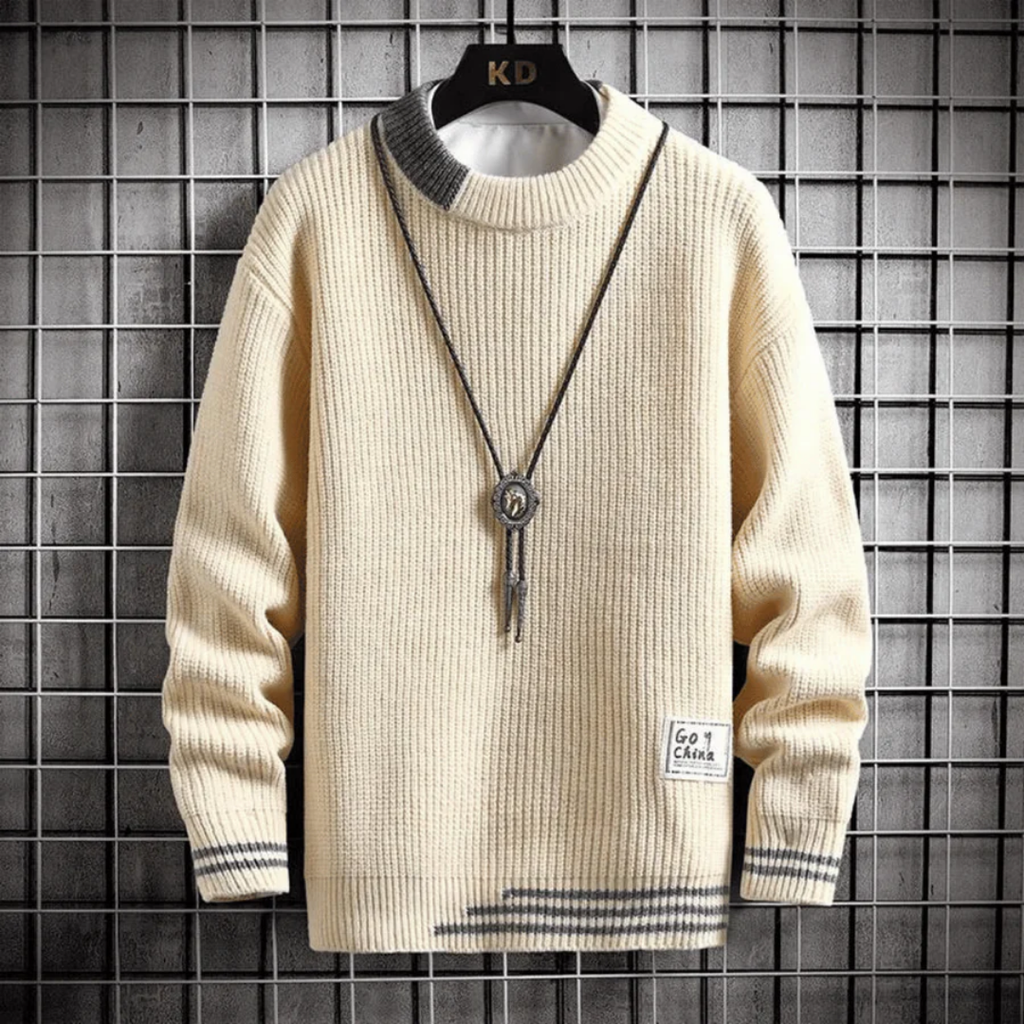 Pull beige en laine avec col rond, détails gris, pendentif long, accroché sur un cintre noir devant un mur en carreaux. Mode automne-hiver tendance.