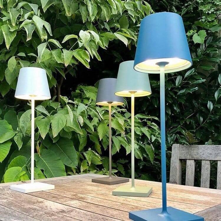 Quatre lampes de table modernes colorées sur une table en bois, entourées de feuillage vert luxuriant dans un jardin. Éclairage extérieur design.