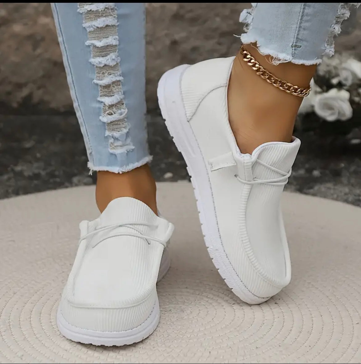Chaussures blanches décontractées pour femmes, portées avec un jean déchiré et un bracelet doré à la cheville, sur fond de tapis beige.