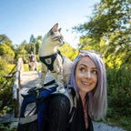 Jeune femme aux cheveux violets portant un chat en harnais sur son dos, marchant dans un parc ensoleillé. Aventure, promenade, nature, complicité.