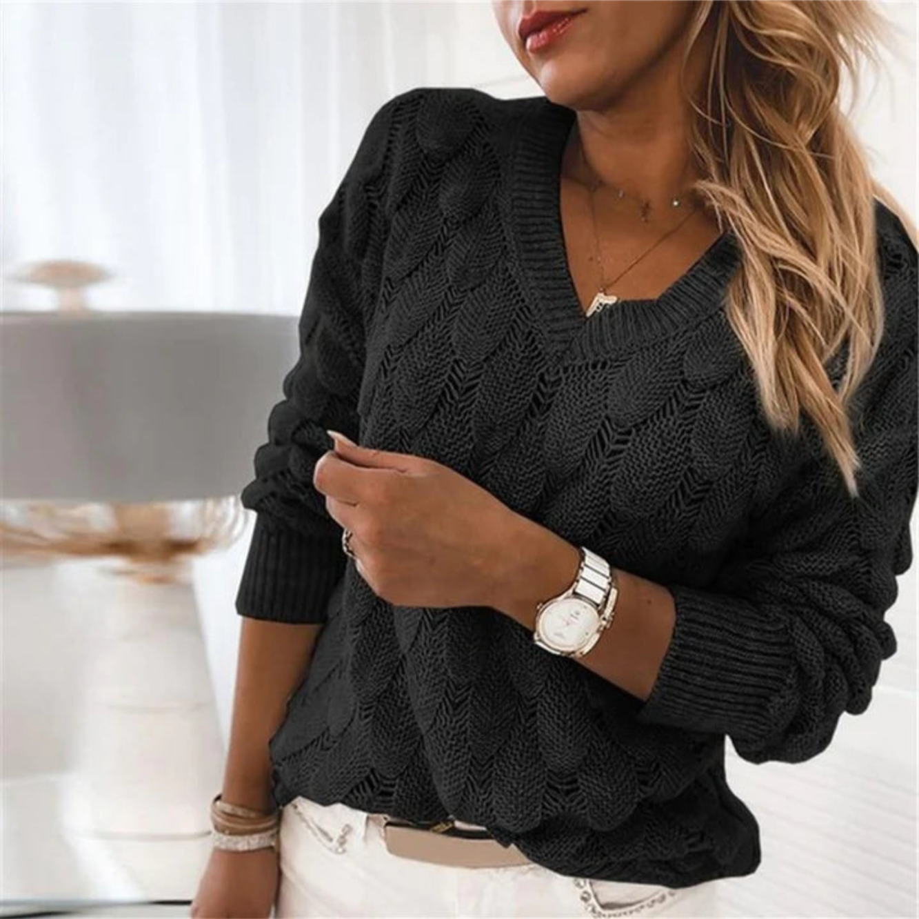 Femme portant un pull noir en tricot torsadé, montre-bracelet blanche, cheveux blonds ondulés, intérieur lumineux. Mode automne-hiver élégante.