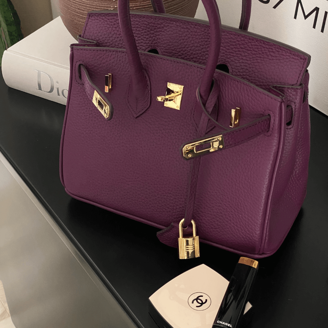 Sac à main en cuir violet avec fermetures dorées, posé sur une table noire, accompagné de maquillage Chanel. Accessoire de luxe élégant.