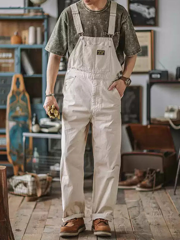 Homme portant une salopette blanche et un t-shirt vert dans un intérieur rustique, avec des accessoires en bois et des étagères en arrière-plan.