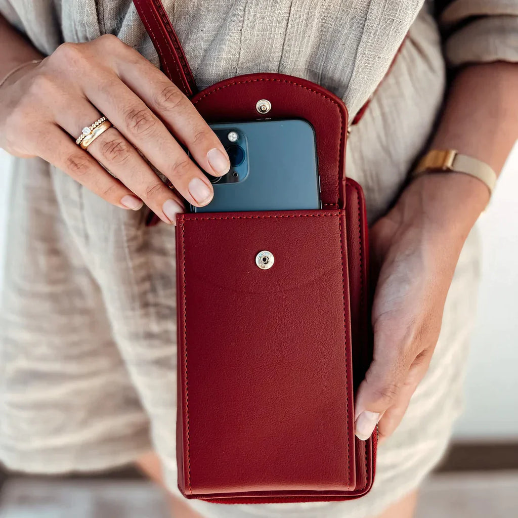 Femme tenant un étui en cuir rouge avec smartphone, robe beige, bague et montre dorée. Accessoire mode pratique et élégant.