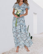 Robe longue fleurie bleue avec manches courtes, femme tenant un bouquet de fleurs blanches, fond minimaliste blanc, élégance estivale.