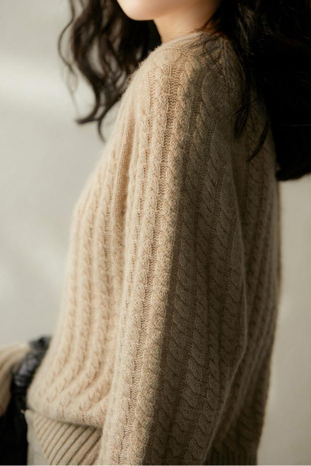 Femme portant un pull en laine torsadée beige, mode automne-hiver, style décontracté et élégant, lumière douce, cheveux noirs ondulés.