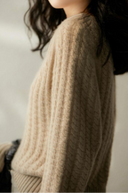 Femme portant un pull en laine torsadée beige, mode automne-hiver, style décontracté et élégant, lumière douce, cheveux noirs ondulés.